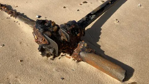 A close up of the rusty remains of World War Two scaffolding poles emerging from sand at Minsmere in Suffolk. It shows a jointed section, with a long pole going off to the left while the pole on the right is broken. At a right angle, a third pole is heading off to the far right corner. It is resting on yellow sand. 