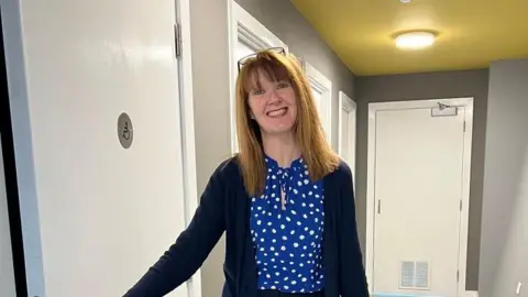 WHC Deanne Shallcross inside the new building The Edge. She is standing by a door with a disabled badge on it and she is smiling. She has light brown hair to her shoulders and a fringe. She is wearing a blue cardigan over a polka dot blue and white blouse.