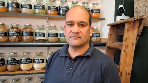 BBC/Seb Cheer Joe Nasson man looks at the camera.He is wearing a navy-blue polo shirt. Behind him, on shelves, a glass jars full of food items.