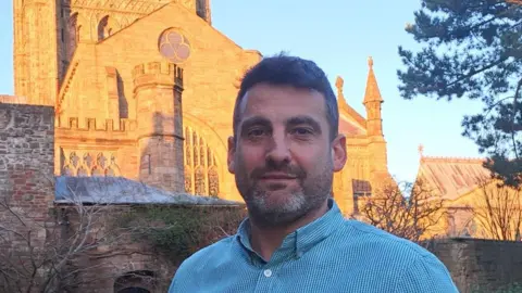 Sam Pratley standing outside Hereford Cathedral. It's a sunny day with a clear blue sky and Mr Pratley is wearing a blue and white striped shirt.