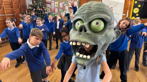 Children in dark blue school uniform pretend to be zombies, chasing a child who is dressed up in a paper machier zombie head. They are in a school hall with a teacher looking on.