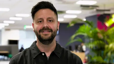 A man stares at the camera. It is a head and shoulder shot. He has dark hair and a beard and moustache and a lip ring. He wears a black shirt. He is in focus while the background is blurred.