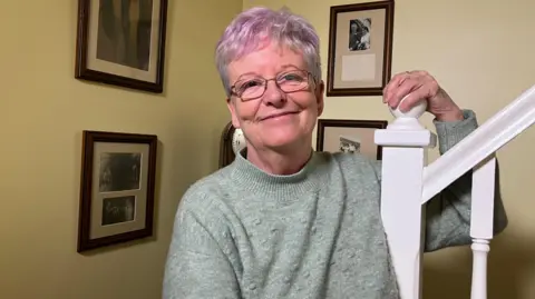 BBC/Victoria Scheer The image shows Maggie standing indoors near a staircase. She is wearing a light green, long-sleeved sweater and has short, light-coloured hair with a hint of purple tone. One hand is resting on the white stair railing, which has a decorative post at the top. The background wall is painted a light yellow shade and features several framed black-and-white photographs arranged in a neat layout. The frames are dark brown and vary in size, with at least four visible in the image.