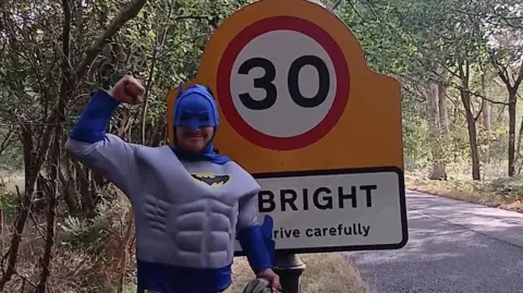 A man dressed a Batman standing next to a road sign.