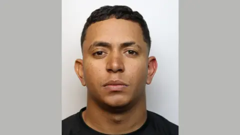 Wiltshire Police A mugshot of a man looking at the camera with a serious expression. He has short black hair and is wearing a black top