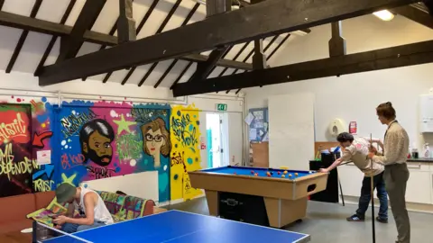 A community room with table tennis and a pool table. A couple of young people are playing pool. Another person is sitting on a sofa. There is a mural on the wall.