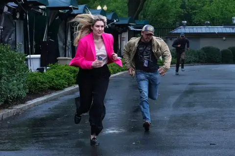 Members of the media clear the North Lawn following reports of gunfire near the White House. A blonde woman is pictured running while wearing a bright pink blazer.