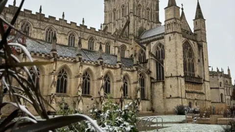 Gloucester Cathedral when it has been snowing. There is snow on the grass and plants surrounding the brick building.