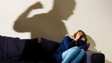 A woman hides her face while sitting on a sofa, while a shadow can be seen on the wall behind of a man raising his fist. 