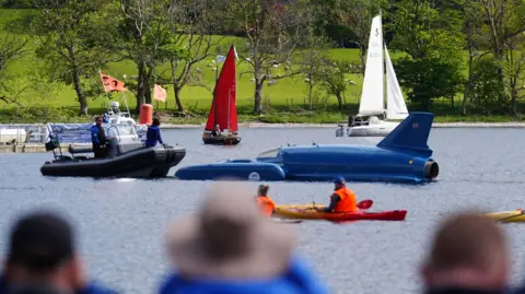 PA Media Bluebird - a cross between a plane and a boat - is towed to its starting point on Coniston Water by a small dinghy. Two other small boats can be seen in the background as people look on from the shore.