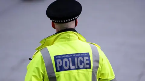 A police officer stands facing away from the camera. He is wearing a high-vis yellow jacket which says 'Greater Manchester Police' on the back. 