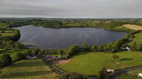 RTE A lake surrounded by green farmland and trees. There's a small carpark in the foreground. 