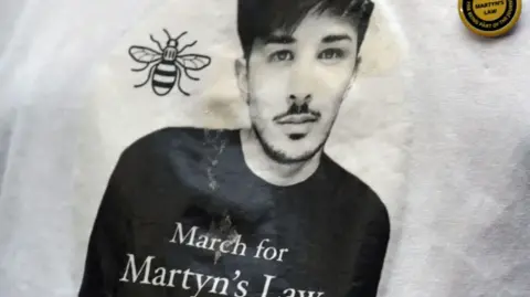 PA Media A black and white image of Martyn Hett, a victim of the Manchester Arena bombing, on a white t-shirt. The bee image is next to Martyn's image