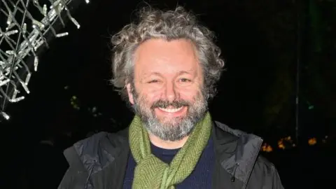 Getty Images Uma cabeça e ombros tiros de Michael Sheen. Ele está do lado de fora e está escuro. Ele tem cabelos curtos cinza, encaracolados e barba cinza e bigode. Ele tem uma jaqueta preta, jumper azul e lenço verde.