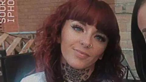 A smiling woman with shoulder length auburn hair. She has two nose piercings and a tattoo on her neck.