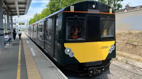 A train has pulled up at a railway station. It is black with windows all down the side and at the front, but there is a bright yellow panel underneath teh driver's window at the front of the train. Some people can be seen waiting for trains on the concrete platform to the left of the image.