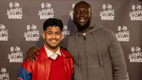 Toby Merritt Stormzy with one arm around Sufiyaan Salam's shoulder. They're smiling and posing in front of a Merky Books-branded backdrop.