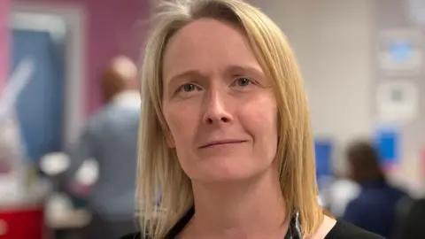 A woman with long blonde hair, a black top and a stethoscope around her neck, smiles slightly as she stands in a hospital.