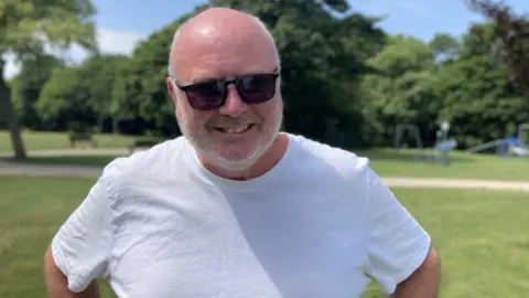 A man is smiling while wearing a white T-shirt and sunglasses. He is standing in a green park field with green tress and a play area behind him.