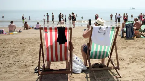 Two people sitting on striped sun loungers on a busy beach on a warm day. Other beach users are wandering around, some are paddling.