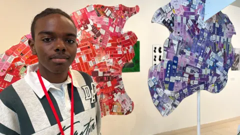 Nicola Haseler/BBC A young man looks into the camera. He is wearing a grey and navy striped polo shirt and a red lanyard. Behind him are two of the pieces which make up the brain sculpture.