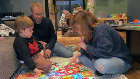 Two adults in dark jumpers and jeans siting on a black and white carpet with their five-year-old son in a black jumper. They are looking at multi-coloured cars on the floor with numbers on them.