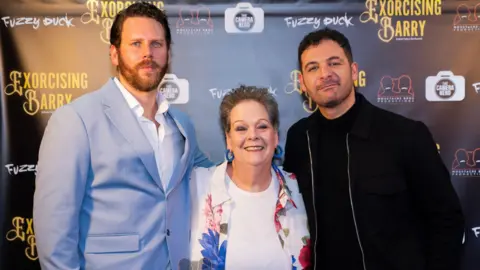 Moustache Bros. Productions Anne Hegerty is smiling at the screening event, to her right is a tall man with a beard and a blue suit. a man dressed in black is on her left. The three have arms around each other.