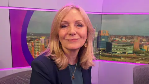 Image shows the Mayor wearing a black suit jacket. She has shoulder length blonde hair and is sitting in a BBC Radio Leeds studio, with an image of the city skyline behind her. 