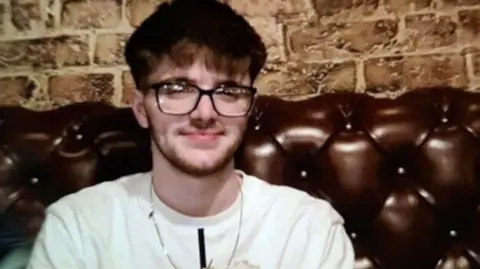 William Webb has dark brown hair and beard and wears a white T-shirt. He sits on a fake leather sofa in front of a brick wall in a bar.