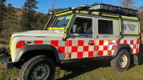 BBC A Patterdale Mountain Rescue vehicle is parked on a grass verge. It is white with a red and white checked pattern on the lower half and Patterdale Mountain Rescue branding.