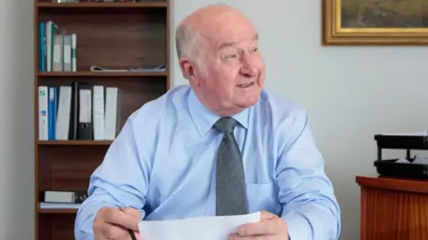 A man wearing a blue shirt and a green tie. He sits at a brown table and there is a bookcase in the background. He is smiling and looking to his left.