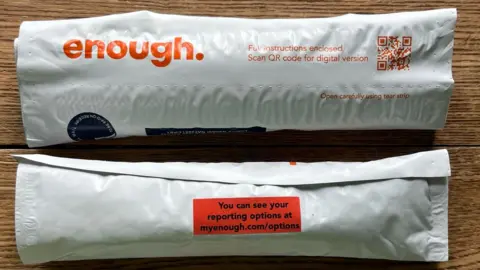 enough Two white narrow packages on a wooden table. The packages are Enough self-swab kits, with the non-profits logo of 'enough' in orange text, a qr instructions code and a red sticker with their website URL.