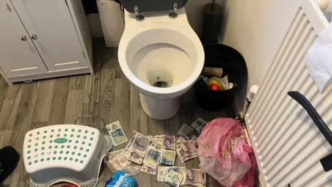 Photograph of banknotes on the floor of a bathroom. The image was taken inside a flat which was raided in Mossley area of Tameside.