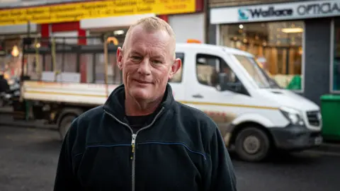 A portrait shot of Darren Ainslie, a light blonde-haired man wearing a black zipper top.Outside, smiles for the camera while behind him a white pick-up truck sits outside an optician.