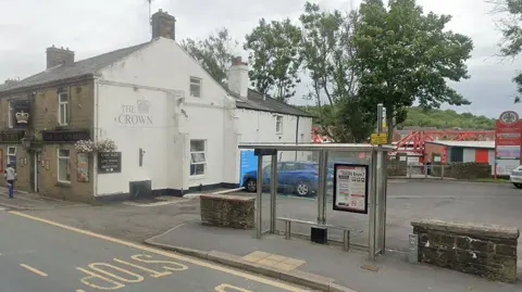 External view of the gable end of the Crown pub in Accrington with a bus stop in front of it and the entrance to Accrington Stanley FC's Wham stadium behind it