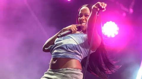 Getty Images Rachel Chinouriri performs on stage. She is holding a microphone stand in one hand and a microphone in the other while biting her finger. She is wearing a blue cropped top and grey skirt. She has long brunette hair. 