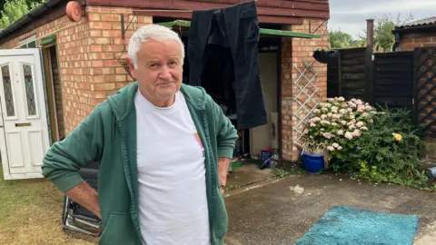 Kate Bradbrook/BBC Imre Berg, de pie al aire libre, en su jardín, junto a un garaje, con las manos en las caderas, con plantas a la derecha y una puerta blanca abierta a la izquierda. Esta puerta de garaje está abierta.