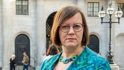 Hillier wears glasses with purple, rounded rims. She has a brown bob and looks sternly at the camera. She is wearing a an aqua and green patchwork blazer with a matching chunky necklace. In the background a grand Portland stone building can be seen and over her left shoulder is a black lamppost with three lamps on top.