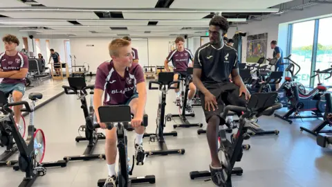Tom Williams/BBC Ranulph (left) and Mo sit on exercise bikes at the club's training facility with other first-team players cycling around them. They are chatting and wearing Norwich City-branded training kit.
