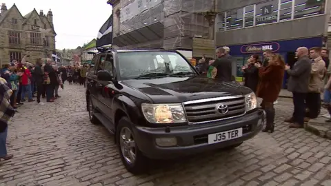 A car drives on the cobbled street in Truro. It has the J35TER number plate. People are gathered on the street to watch.
