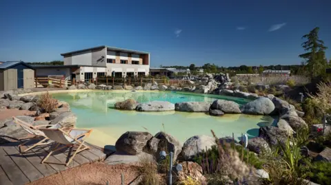 Love2Stay A small shallow lake surrounded by large stones, sand, grass and an area of decking with a large flat-roofed building on the far side