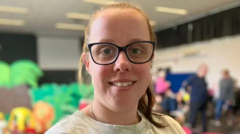 A woman with glasses and her hair tied back. She is smiling and looking into the camera. behind her are parents and children.