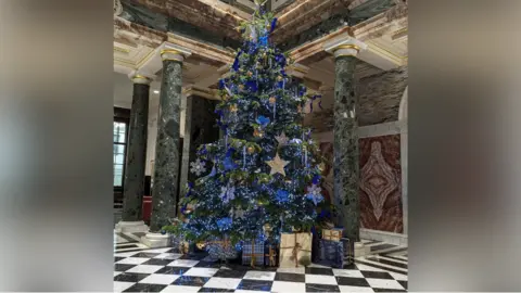 A large Christmas tree with blue decorations inside a marble hall. 