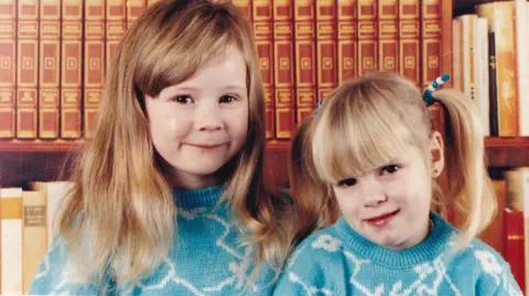 Family of Kirsty Wilkinson Hayley and Kirsty Wilkinson are two white girls aged approximately five and three. Hayley, left, has chest length blonde hair with a long fringe brushed to the side. Kirsty has blonde hair worn in two bunches. They are wearing identical turquoise jumpers with irregular white lines forming patterns on it. They are both smiling at the camera standing in front of bookshelves.