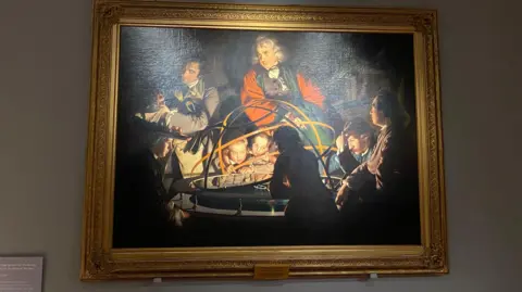 The picture features a painting of a group of people studying a contraption exploring the solar system.