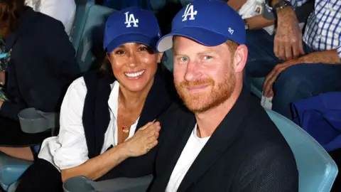 Meghan and Harry in blue baseball caps watching a game at the Dodgers Stadium in Los Angeles