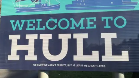 A blue and turquoise sign which reads Welcome to Hull with We know we aren't perfectm but at least we aren't Leeds below in smaller writing
