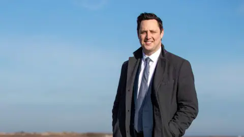 Ben Houchen is smiling into the sun wearing a grey overcoat on top of a navy suit. He is smiling and a flat stretch of land can be seen behind him.