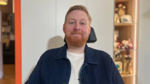 A man with red hair and beard sitting smiling in a wheelchair inside a home. He is wearing a denim jacket over a white t-shirt.
