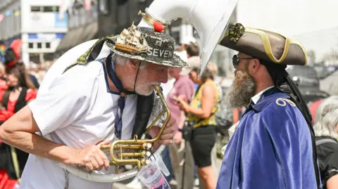 Hugh Hastings A street performer wearing a decorated hat and carrying a large brass instrument plays closely in front of another costumed performer in a blue cloak and tricorn-style hat. The busy festival street behind them is filled with people, colourful outfits and buildings, capturing a lively parade atmosphere in bright daylight.